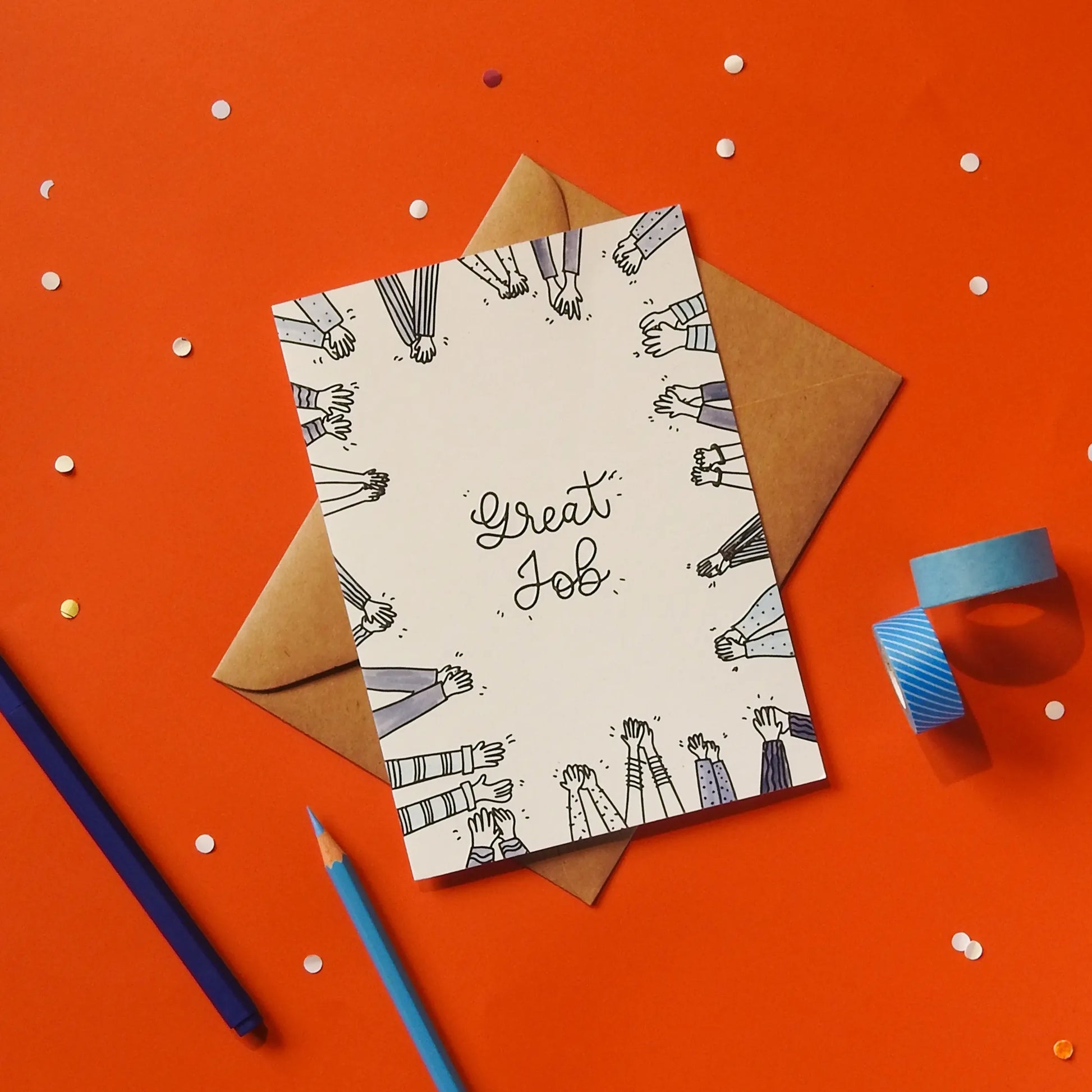 A white greeting card with 'Great Job' text and hand illustrations, accompanied by a brown envelope against a red background with stationery items.