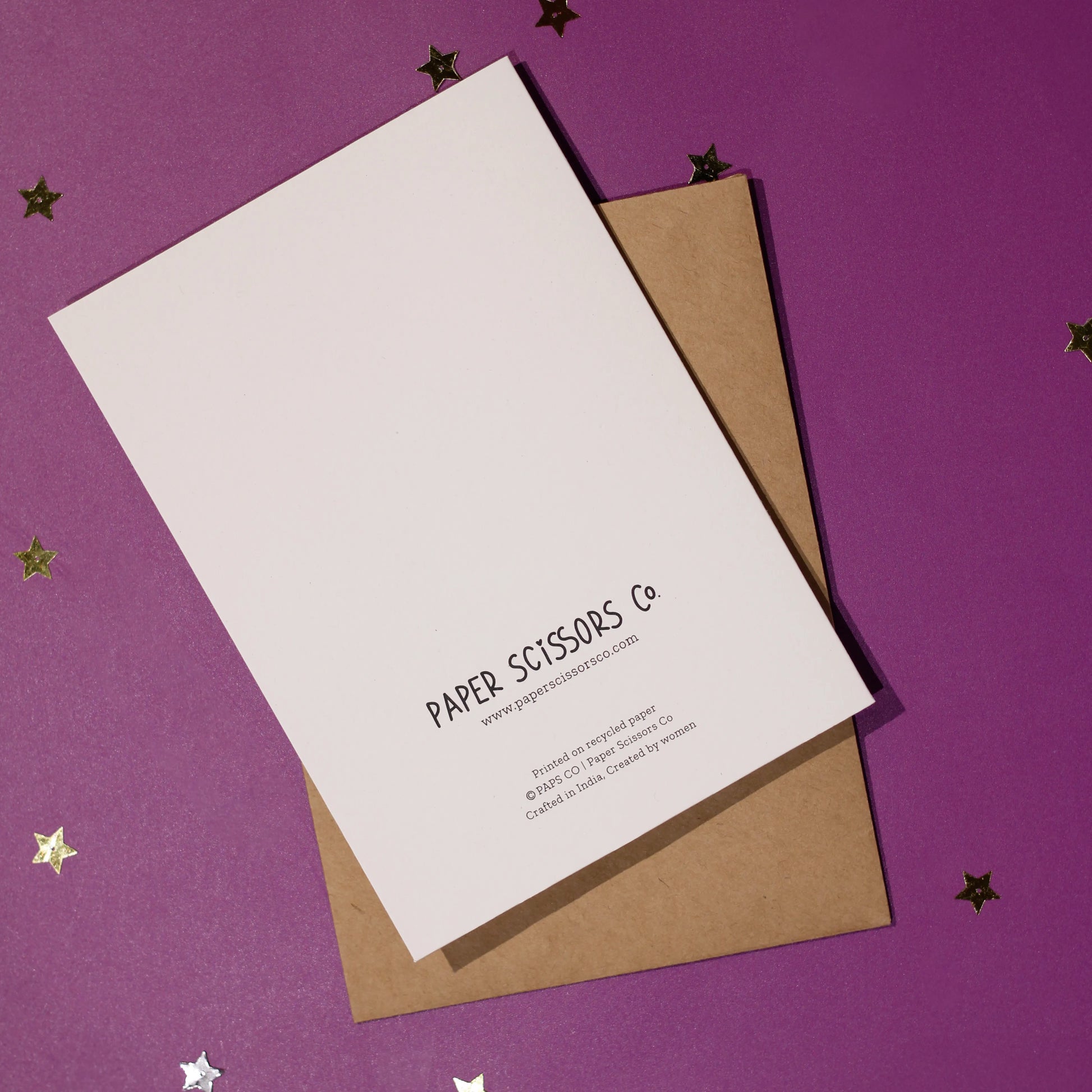 A white greeting card back cover highlighting Paper Scissors Co. accompanied by a brown envelope with decorative elements on a purple background.