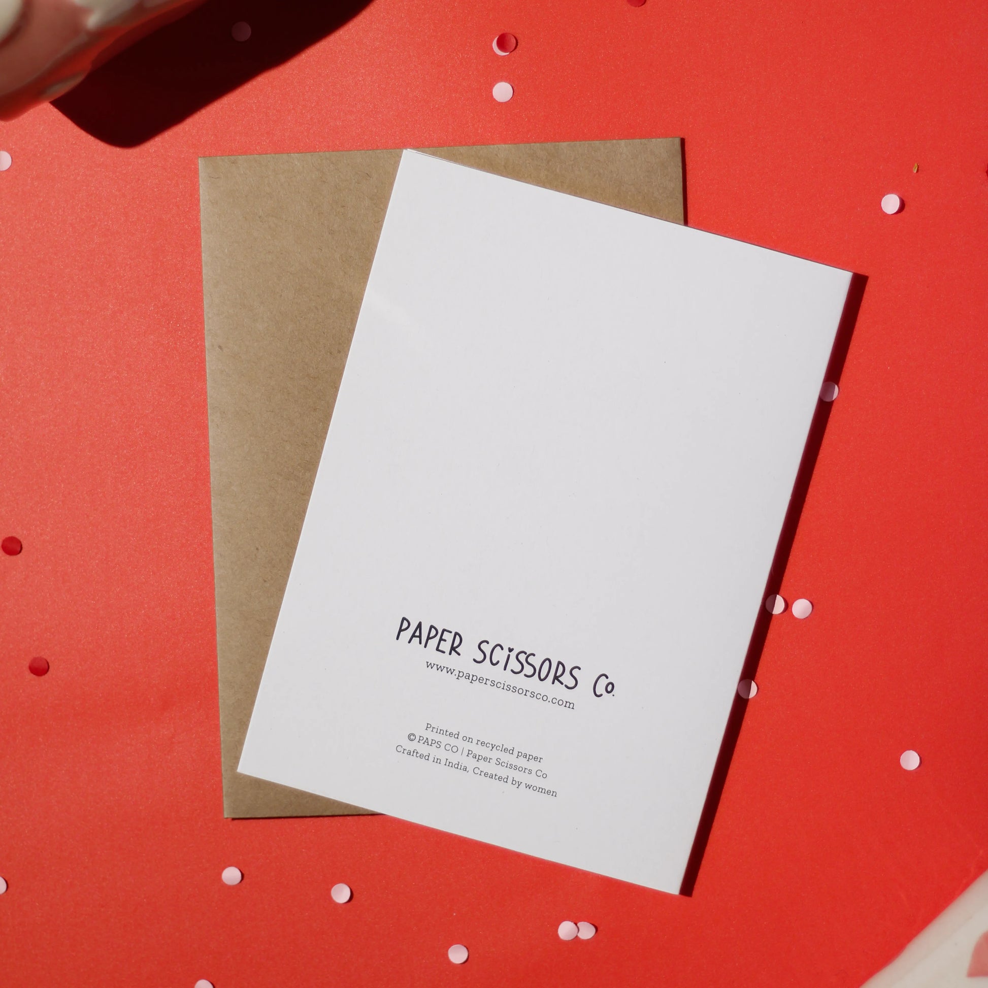 A white greeting card with 'Paper Scissors Co.' accompanied by a brown envelope text against a red background with decorative elements.
