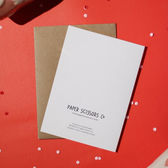 A white greeting card with 'Paper Scissors Co.' accompanied by a brown envelope text against a red background with decorative elements.