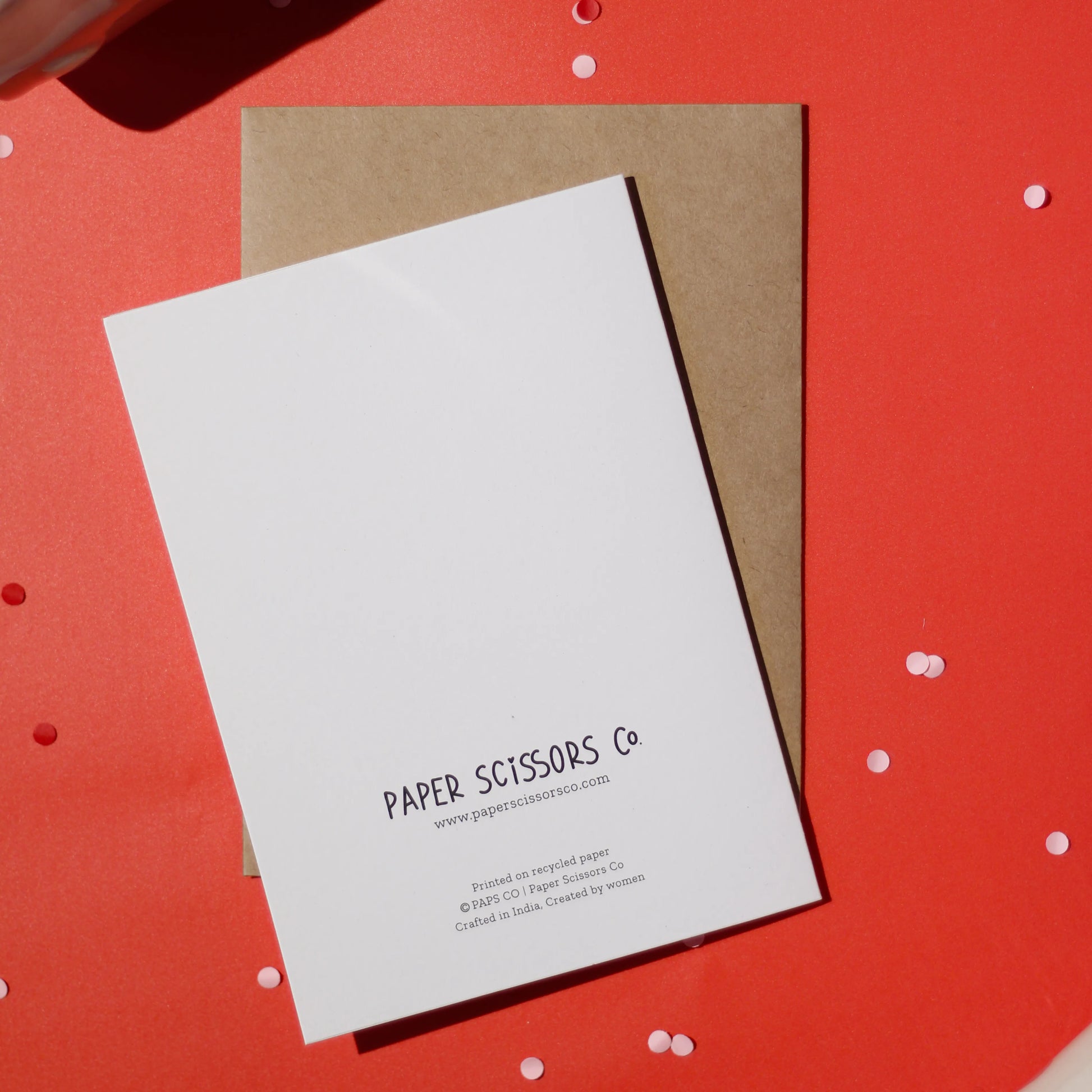 A white greeting card with 'Paper Scissors Co.' branding accompanied by a brown envelope against a red background with confetti.