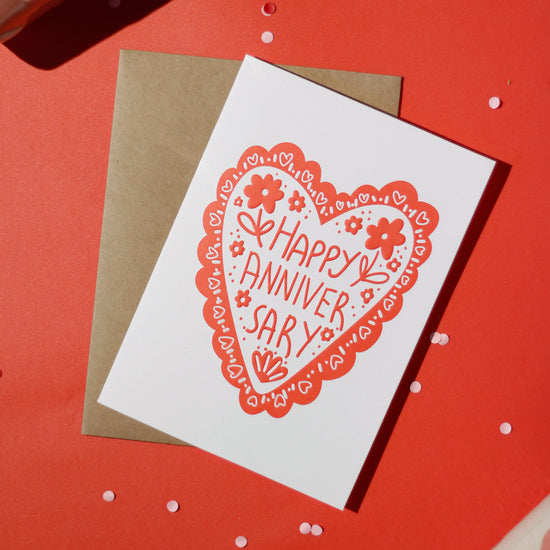 A white greeting card with 'Happy Anniversary' text and an illustrated heart, accompanied by a brown envelope on a red background with decorative elements.