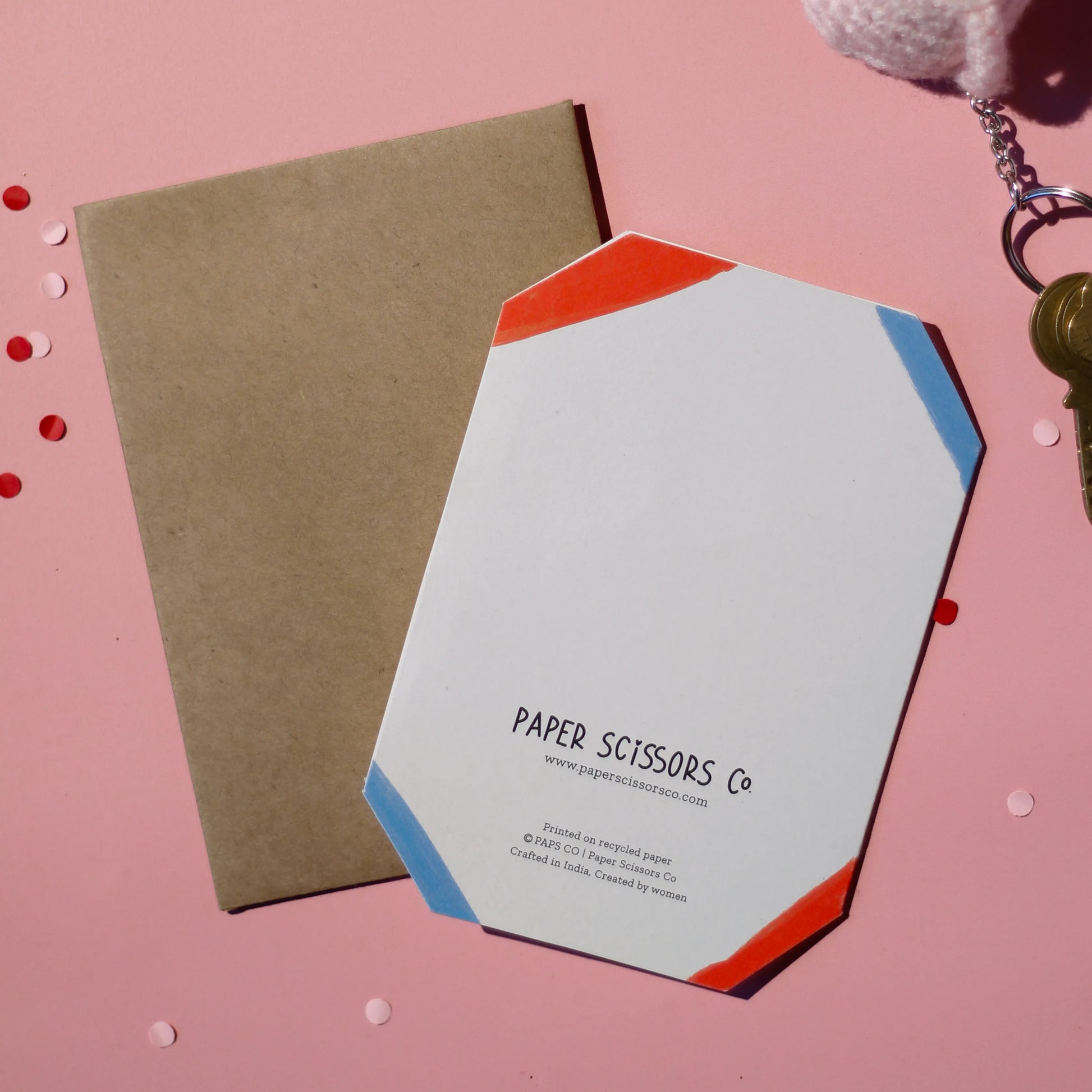 A hexagonal die-cut card with 'Paper Scissors Co.' branding accompanied by a brown envelope on a light pink background.