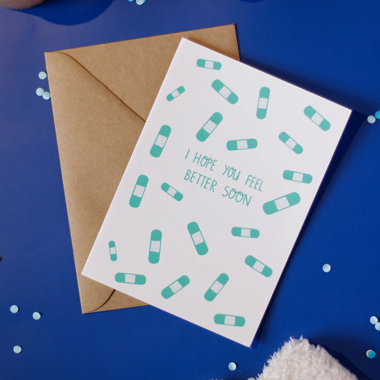 A white greeting card with bandage pattern and 'I hope you feel better soon' text on a dark blue background with decorative elements.