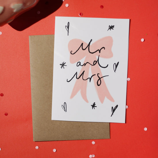 A white greeting card with 'Mr and Mrs' text in black embossment and a pink bow illustration, accompanied by a brown envelope on a red background with decorative elements.