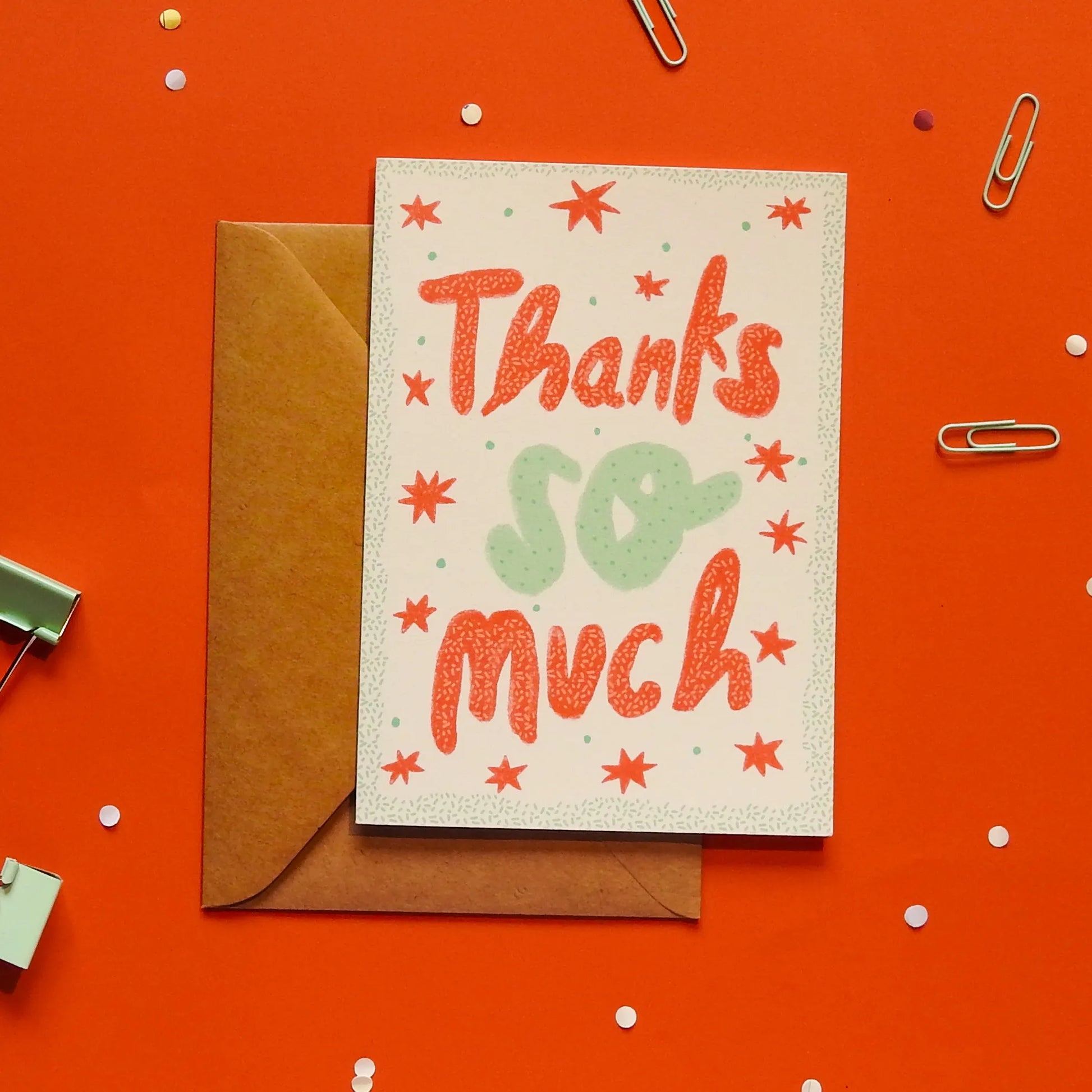 A white greeting card with 'Thanks so much' text, with a brown envelope on an orange background with decorative elements.