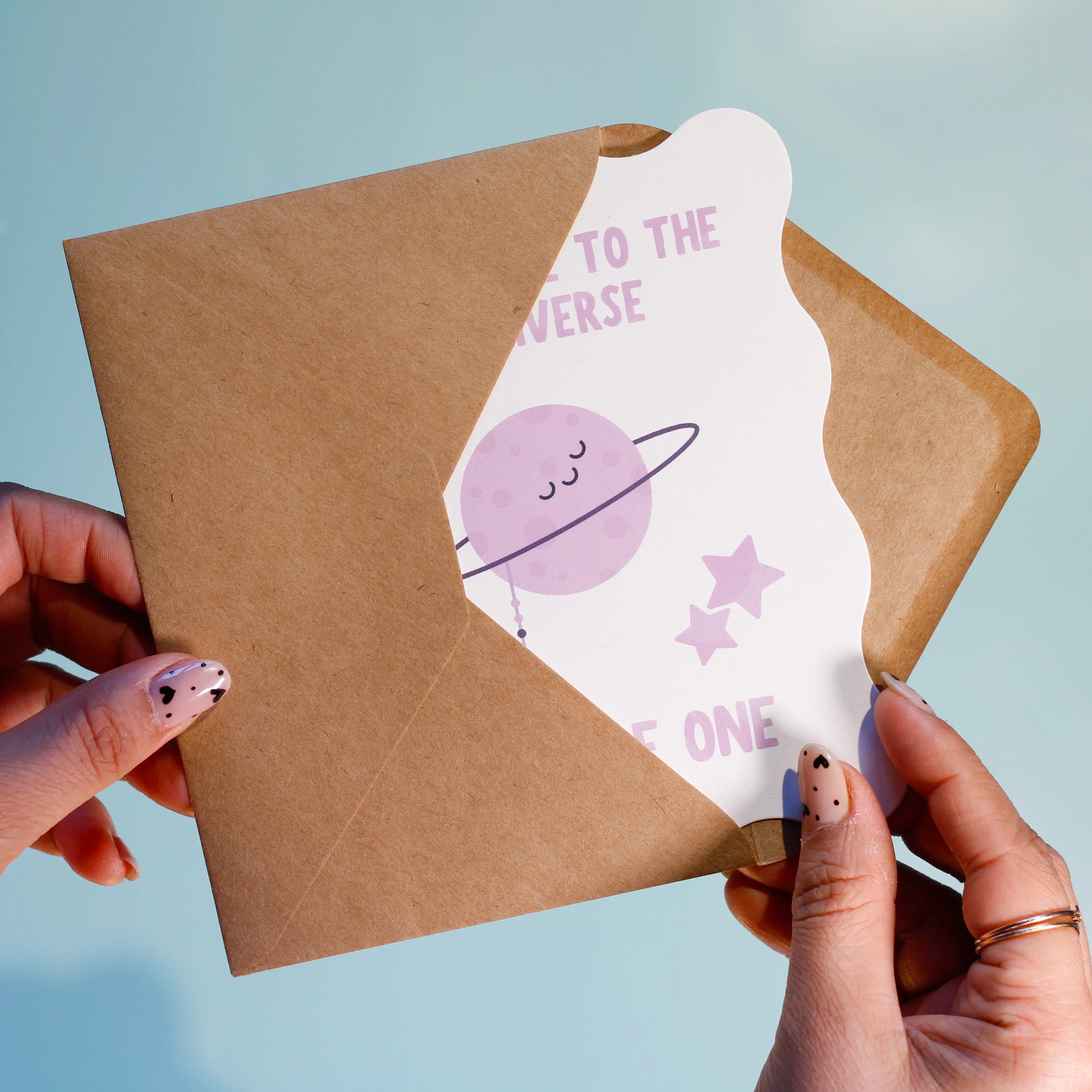 A person holding a brown envelope with "Welcome to the universe, Little One" text, featuring a purple planet and star design on a light blue background.