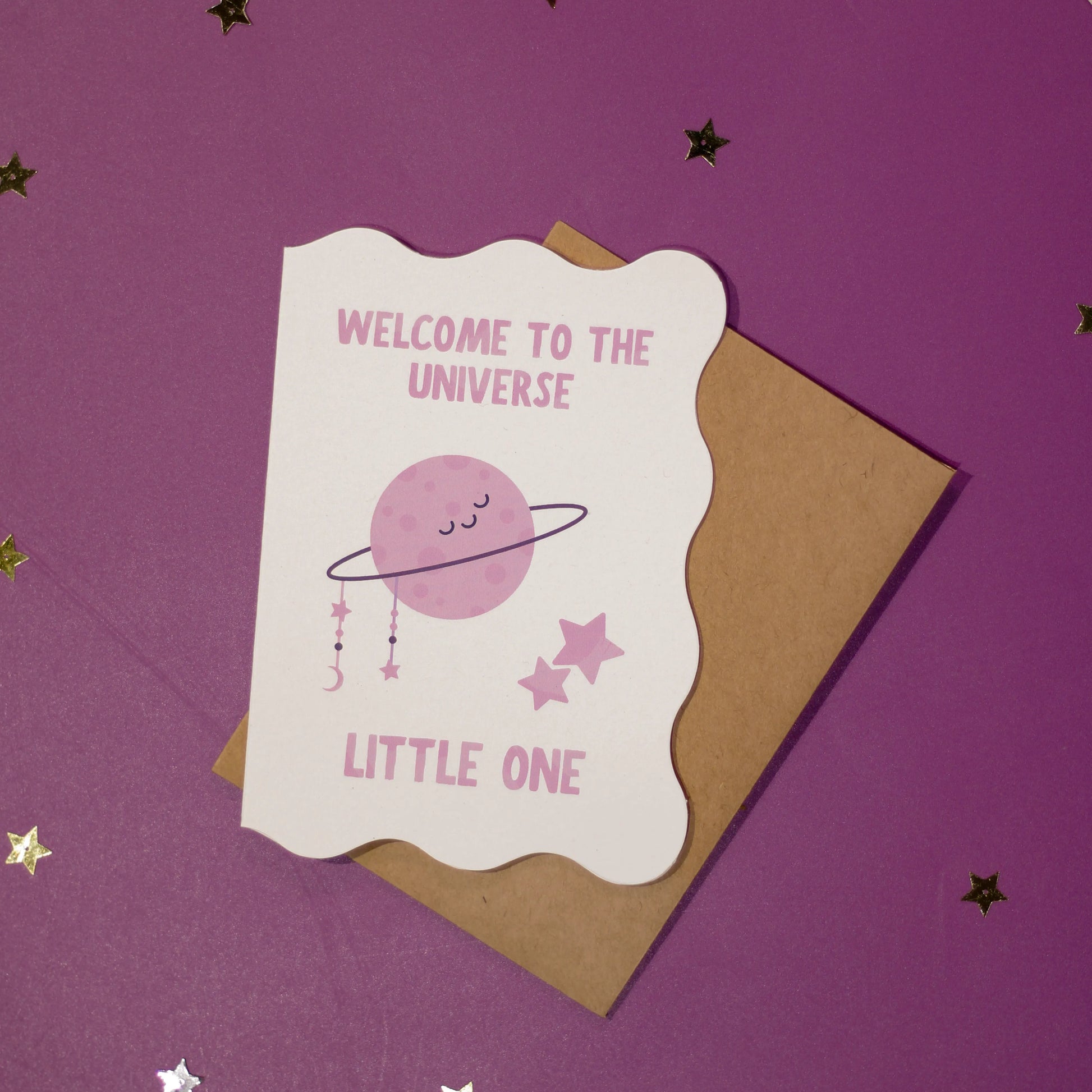 A white die-cut greeting card with 'Welcome to the Universe Little One' text and space-themed illustration on a purple background with decorative elements.