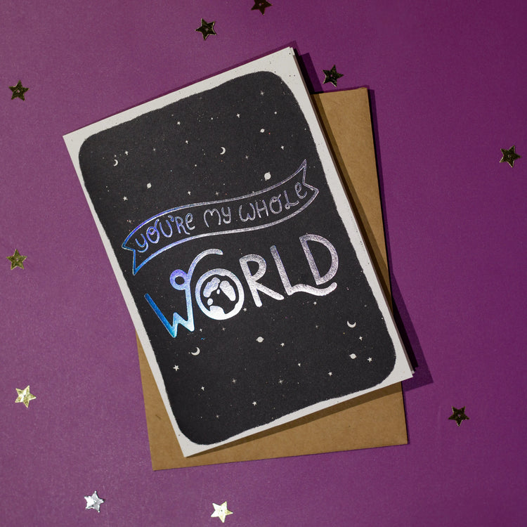 Greeting card with 'You're my whole world' in silver holographic effect, accompanied by a brown envelope on a purple background with decorative elements.