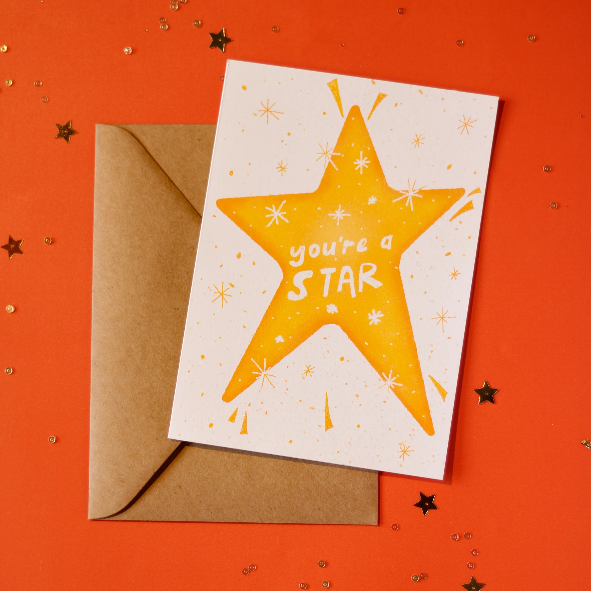 A white greeting card with a yellow star illustration and 'you're a star' text, accompanied by a brown envelope against an orange background with decorative elements.