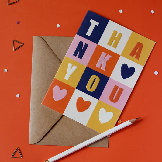 A 'Thank You' greeting card with colour blocks and hearts, with a brown envelope on an orange background with decorative elements.
