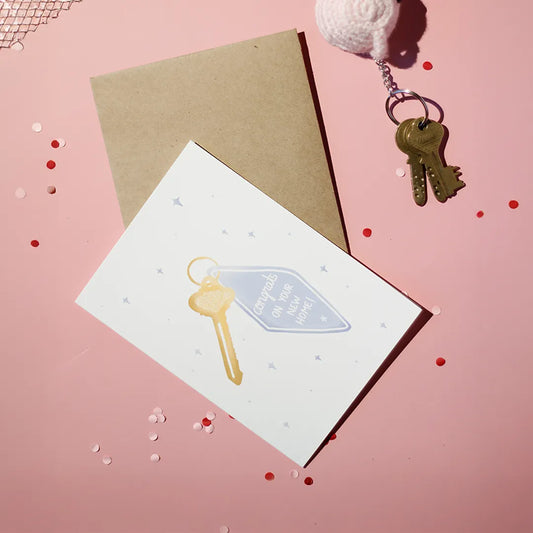 A white greeting card with a key illustration, with text 'Congrats on Your New Home', accompanied by a brown envelope against a light pink background with decorative elements.