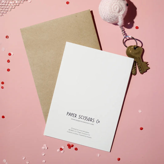 A white greeting card with 'Paper Scissors Co.' text accompanied by a brown envelope on a light pink background with a brown envelope and decorative elements.