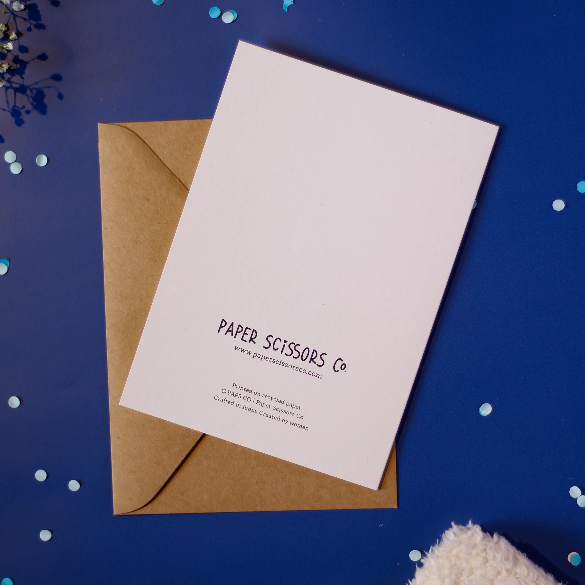 A white greeting card back cover featuring 'Paper Scissors Co.' branding, accompanied by a brown envelope on a dark blue background with decorative elements.