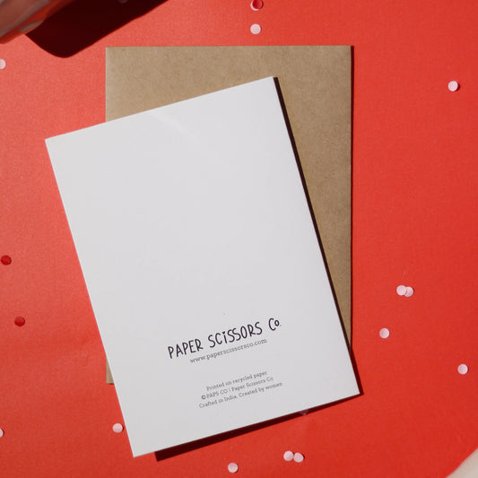 A white greeting card with 'Paper Scissors Co.' branding accompanied by a brown envelope against a red background with confetti.