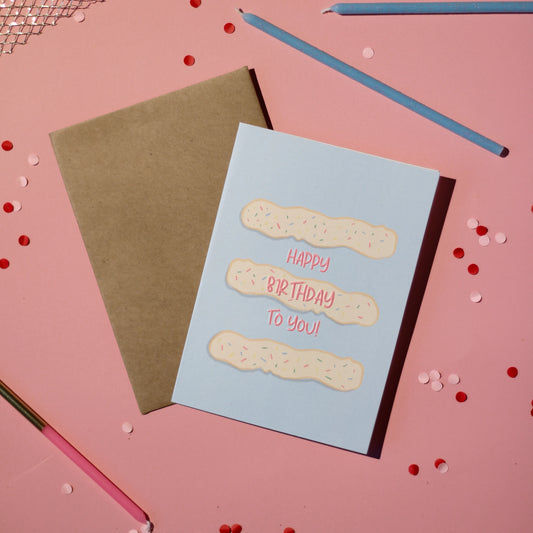 A blue card with frosting and 'Happy Birthday To You!' text on a light pink background with confetti.