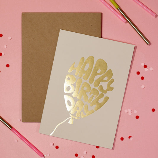 A white greeting card with a gold foiled Happy Birthday text in a balloon with a brown envelope on a light pink background with decorative elements. 