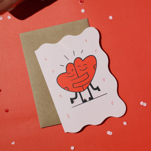 A die-cut greeting card with two illustrated hearts hugging, accompanied by a brown envelope on a red background with decorative elements.