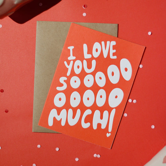 A red greeting card with 'I love you sooooo much!' text accompanied by a brown envelope on a red background with decorative elements.