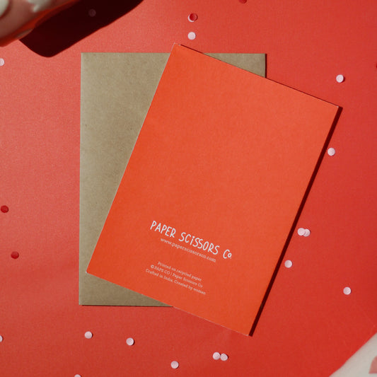 A red greeting card with 'Paper Scissors Co.' text accompanied by a brown envelope on a red background with decorative elements.
