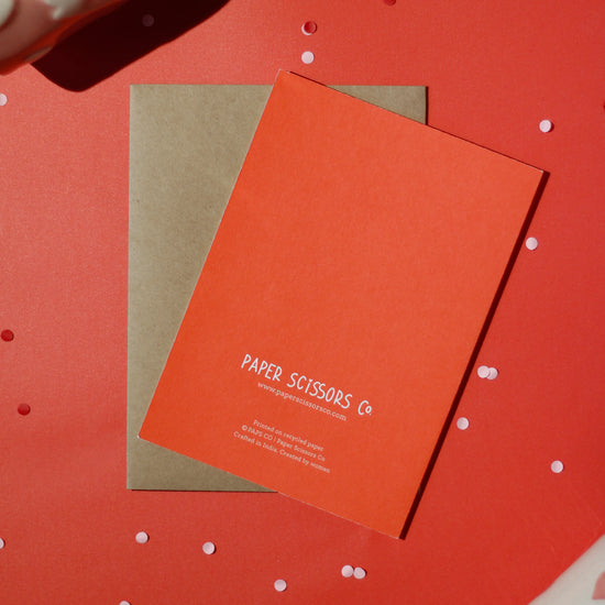 A red greeting card with 'Paper Scissors Co.' text accompanied by a brown envelope on a red background with decorative elements.
