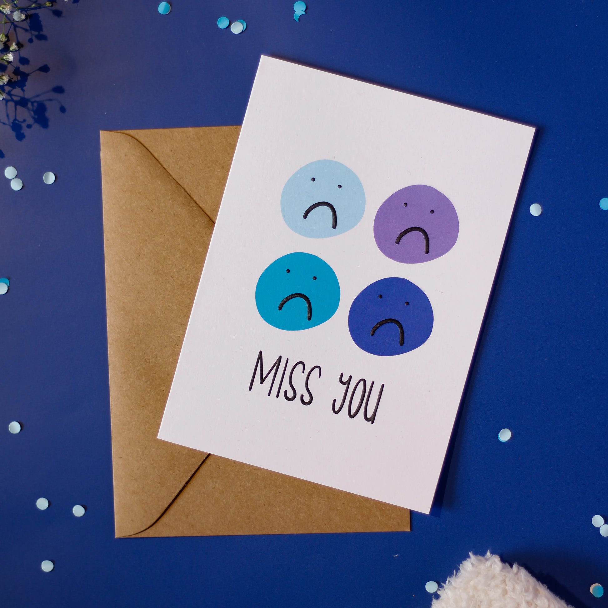 'Miss You Sad Emojis' Greeting Card with a brown envelope on a dark blue background with decorative elements.