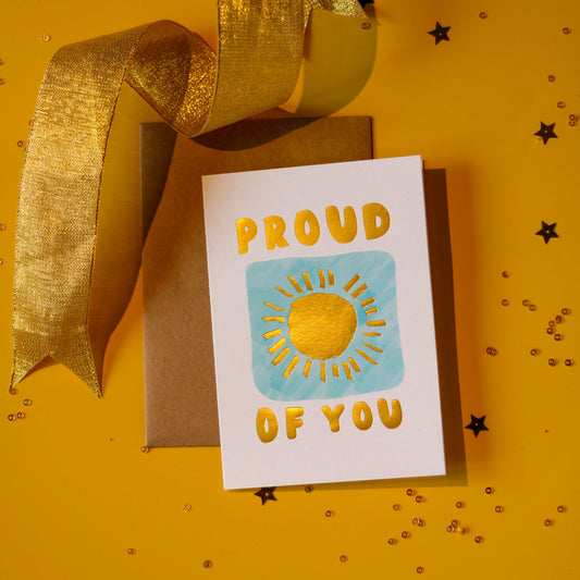 A white greeting card with 'Proud of You' text and sun design in gold foil on a blue illustrated square against a yellow background with a gold ribbon and decorative elements.