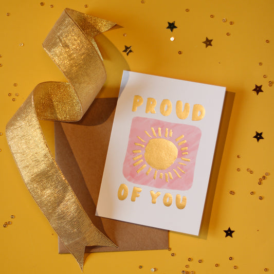 A white greeting card with 'Proud of You' text and sun design in gold foil on a pink illustrated square against a yellow background with a gold ribbon and decorative elements.