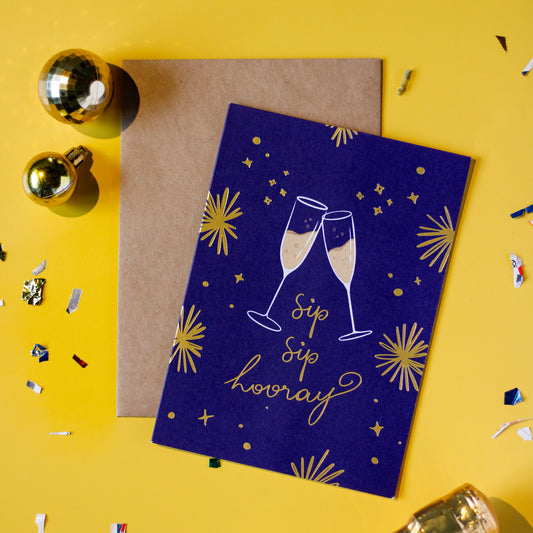 A dark blue greeting card with champagne glasses and 'Sip Sip Hooray' text with a brown envelope on a yellow background with decorative elements.