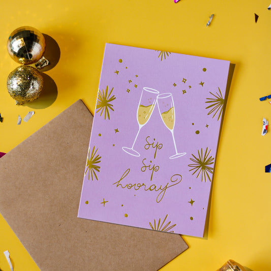 A light purple card with champagne glasses and 'Sip Sip Hooray' text with a brown envelope on a yellow background with decorative elements.