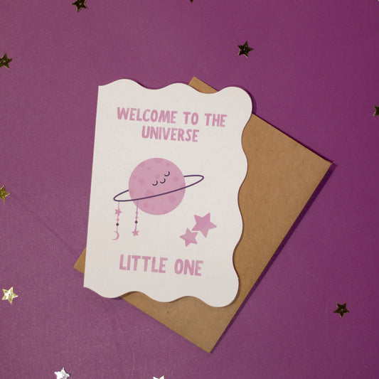 A white die-cut greeting card with 'Welcome to the Universe Little One' text and space-themed illustration on a purple background with decorative elements.