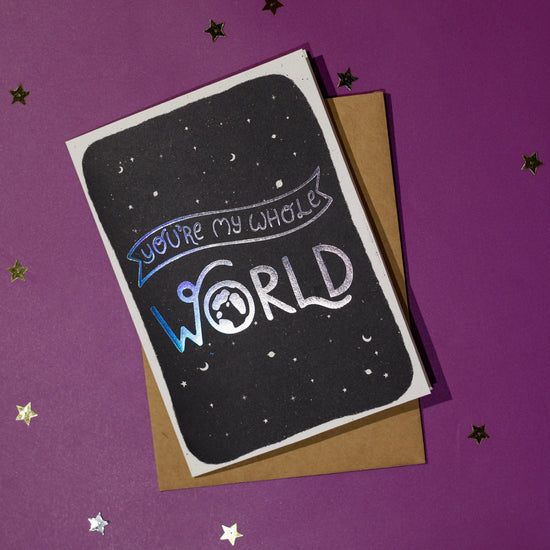Greeting card with 'You're my whole world' in silver holographic effect, accompanied by a brown envelope on a purple background with decorative elements.
