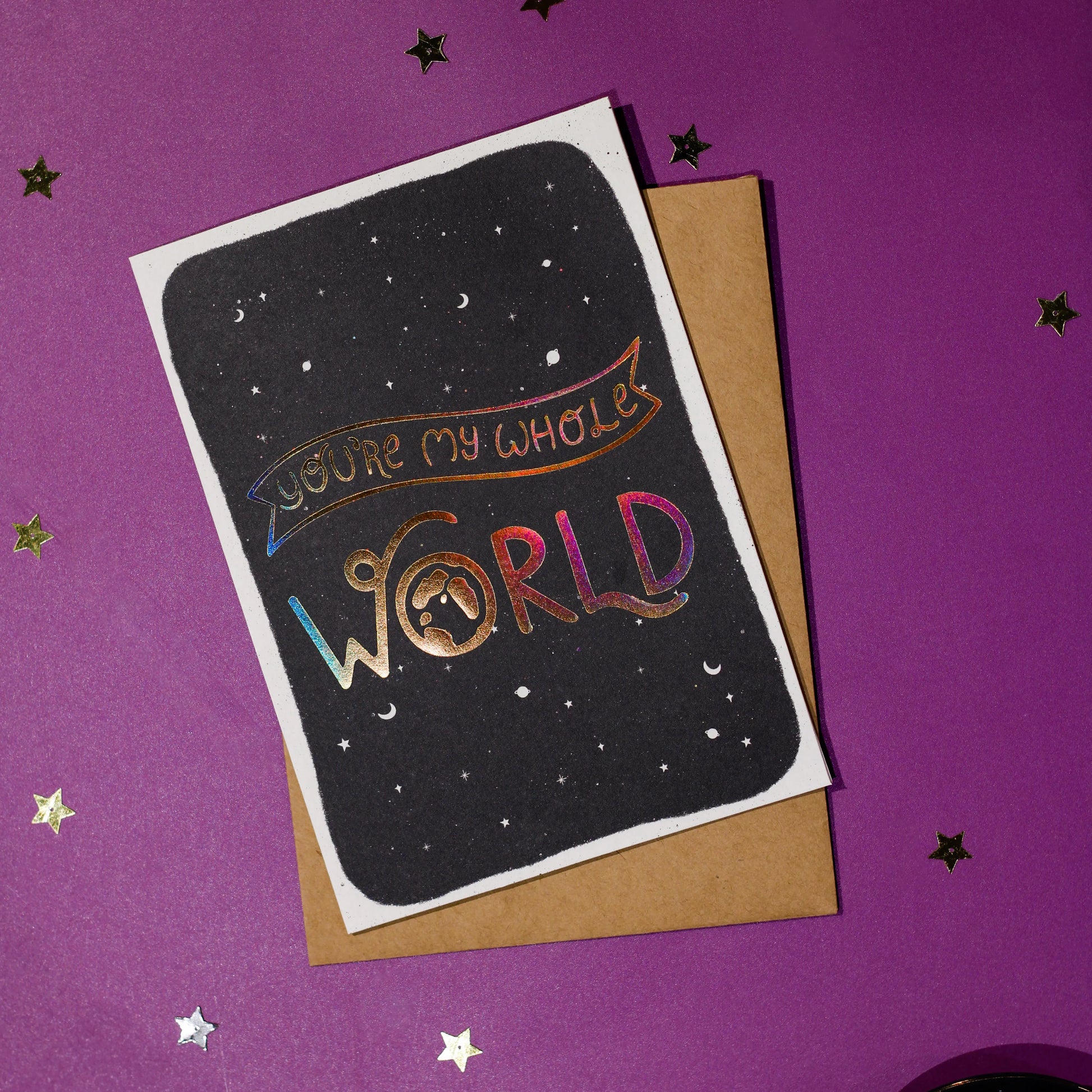 Greeting card with 'You're my whole world' in gold holographic effect, accompanied by a brown envelope on a purple background with decorative elements.