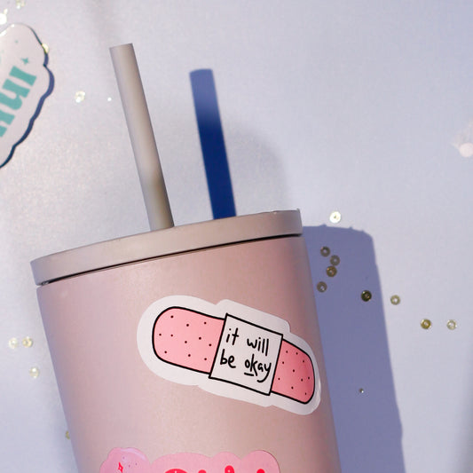 a cute pink tumbler with our 'it will be okay' sticker on a light blue background.